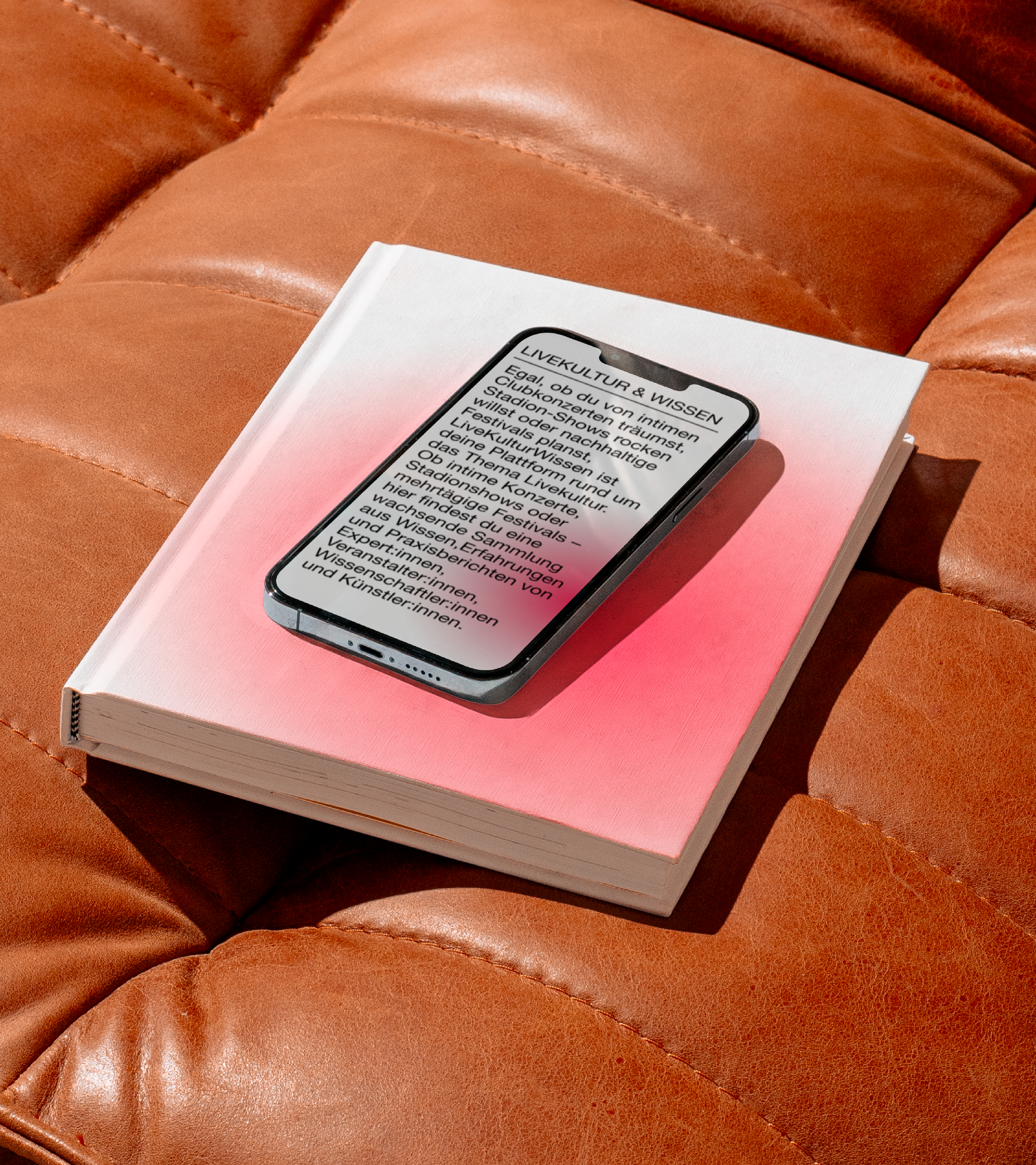Datenbank für LiveKulturWissen Ein Handy liegt auf einem weiß-pinken Buch, beide Elemente liegen auf einer braunen Ledercouch.