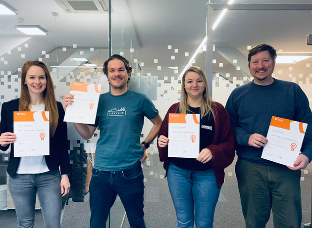 Four people stand indoors, each holding a typo3 certificate, smiling at the camera. The background features a modern office setting.