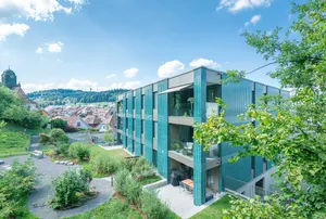 Mehrfamilienhaus in Willisau mit grüner Keramikfassade, vertikal strukturierten Elementen und großen Balkonen, umgeben von Grünflächen und Kieswegen.