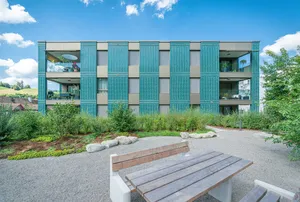Residential building in Willisau featuring a green ceramic façade with vertical elements and large balconies, surrounded by greenery and gravel paths.