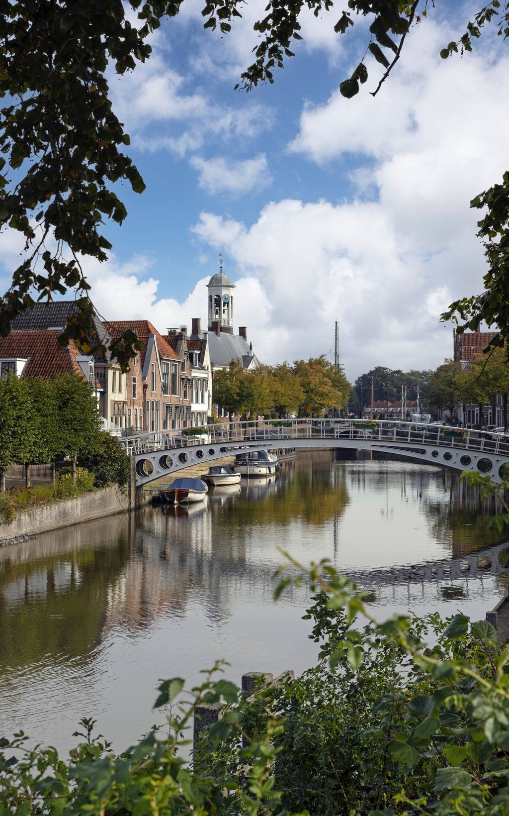 Ein ruhiger Kanal in einer malerischen Stadt mit einer weißen Brücke, Booten und historischen Gebäuden unter einem teilweise bewölkten Himmel, umrahmt von belaubten Ästen.