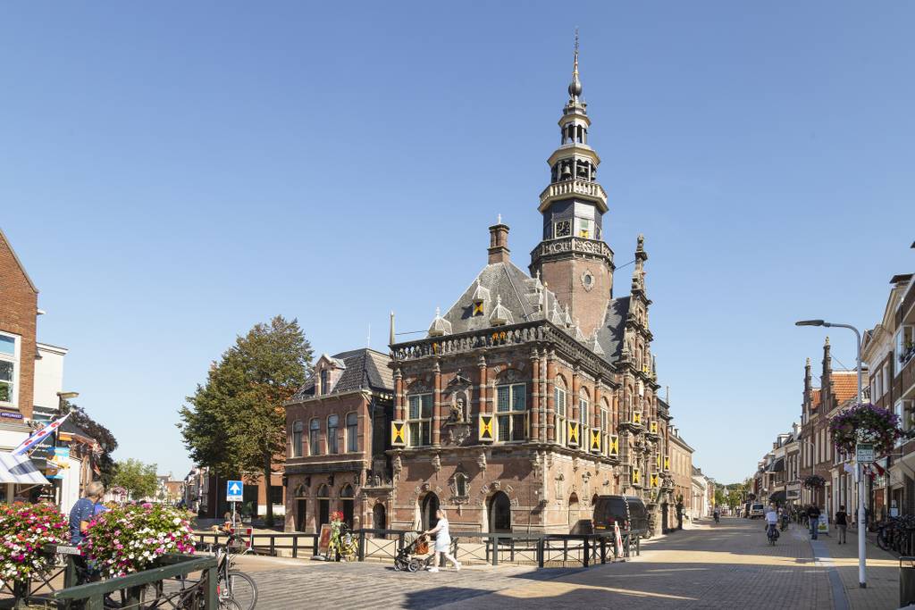 Historisches Backsteingebäude mit einem hohen Uhrenturm und kunstvollen Details, an einer Kopfsteinpflasterstraße unter blauem Himmel gelegen.