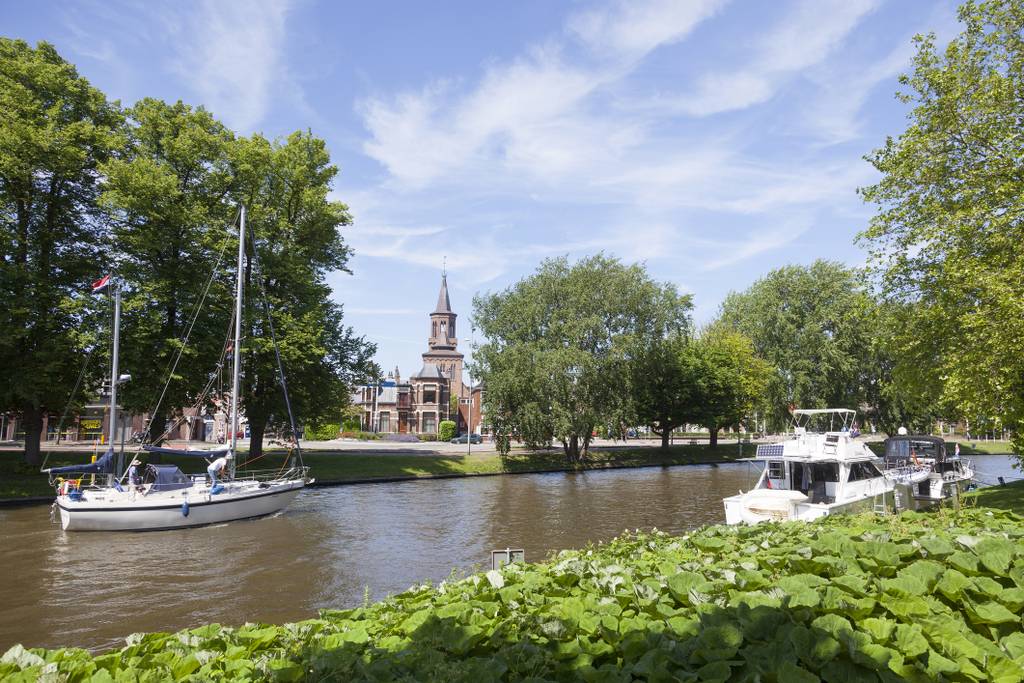 Boote auf einem Kanal mit üppigem Grün und einem Kirchturm im Hintergrund unter einem blauen Himmel.