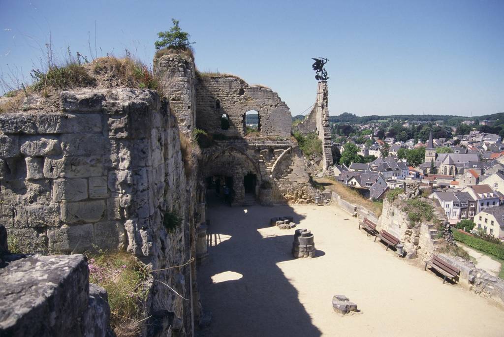 Ruinen einer Steinburg mit Bogenfenstern, die eine Stadt und die Landschaft unter einem klaren blauen Himmel überblicken. Bänke säumen den sandigen Innenhof.