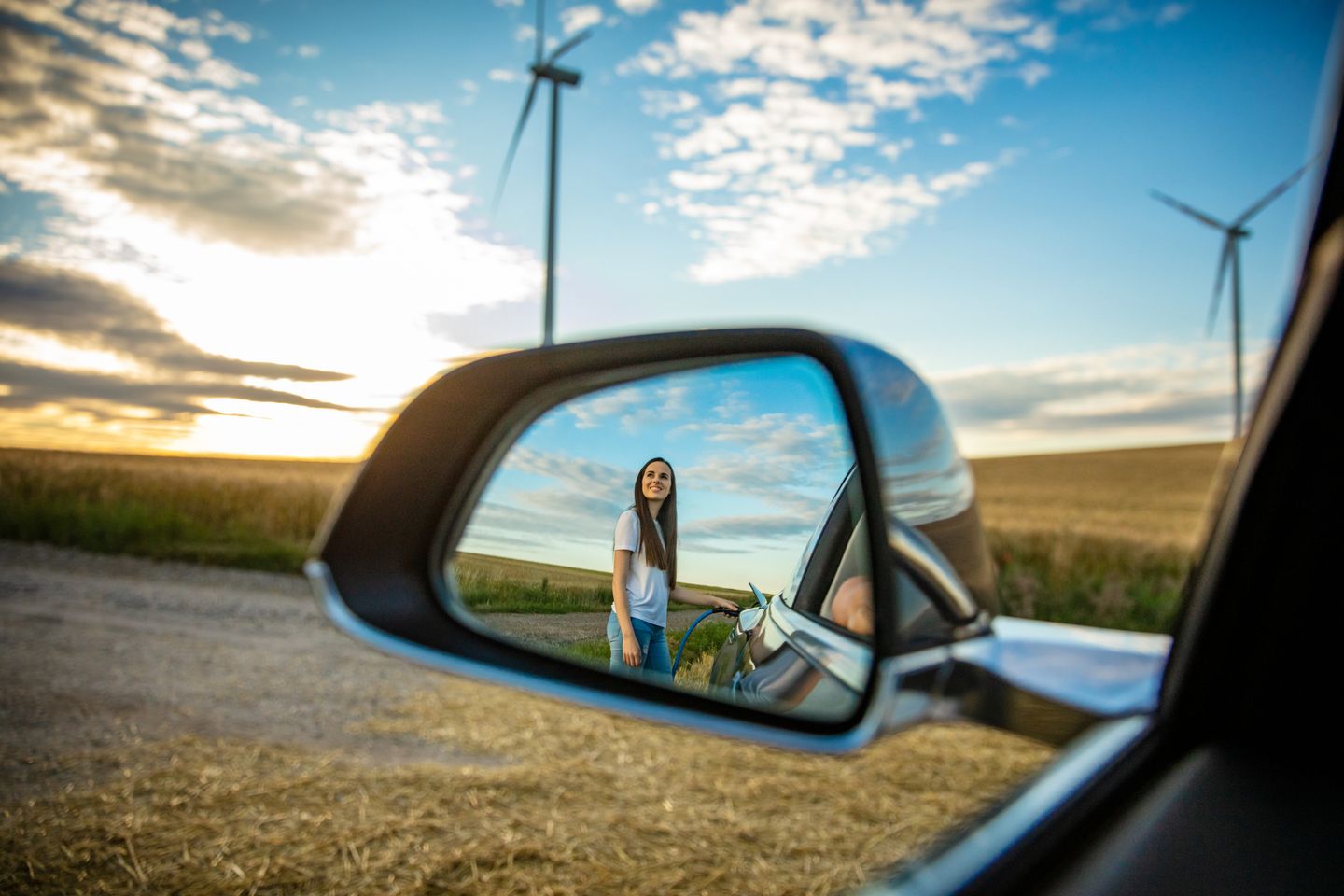 Reflexion einer jungen Frau im Außenspiegel beim Laden eines Elektroautos in einem ländlichen Gebiet mit Windrädern im Hintergrund.