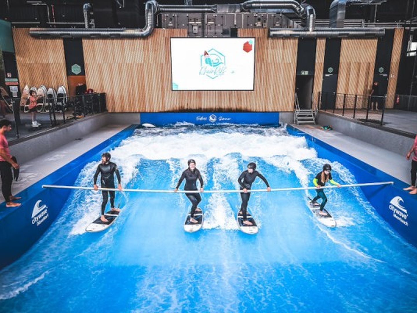 Surf-Schüler:innen stehen auf ihren Surfbrettern im Wavepool des RheinRiffs und halten sich an einer Stange fest. © RheinRiff