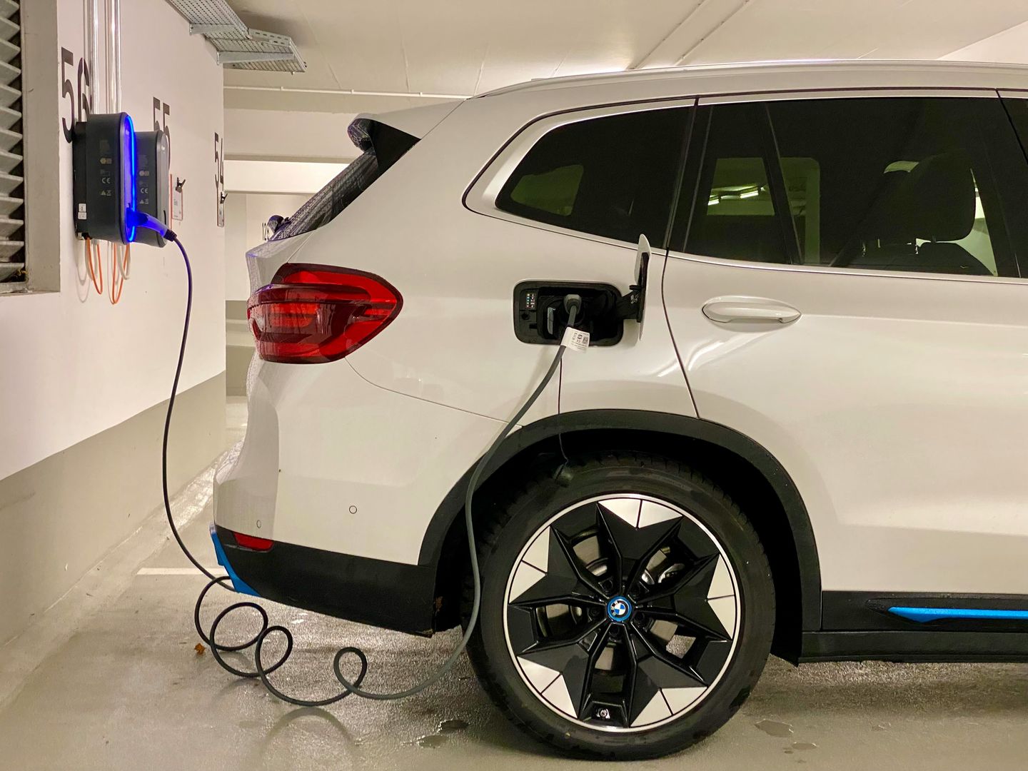Ein weißes Elektroauto lädt in einer Garage an einer Wandladestation; das Ladekabel ist am hinteren Anschluss des Fahrzeugs eingesteckt. Foto von Eren Goldman auf Unsplash
