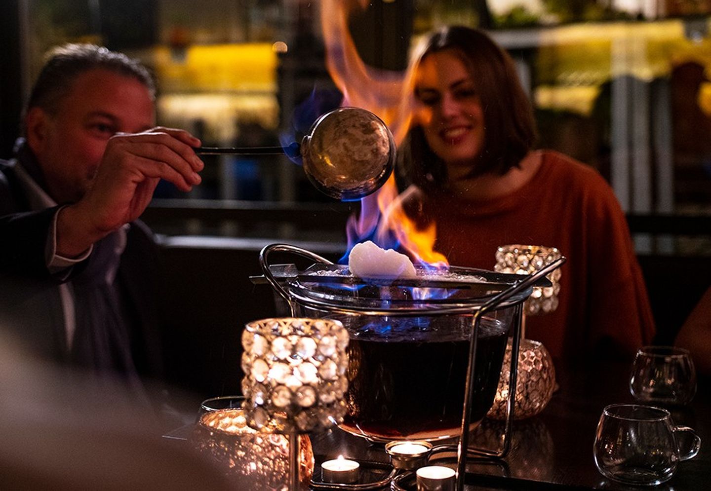 Feuerzangenbowle wird von einem Mann mit einer Kelle mit Rum übergossen, während aus dem Zuckerhut hohe Flammen schlagen.