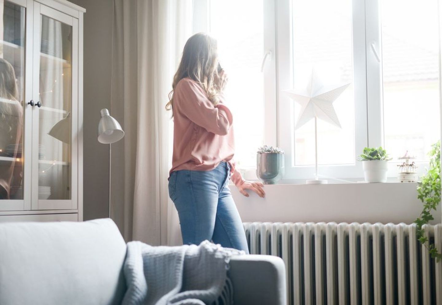 Eine Frau steht beim Telefonieren an einer Heizung und schaut aus dem Fenster. 