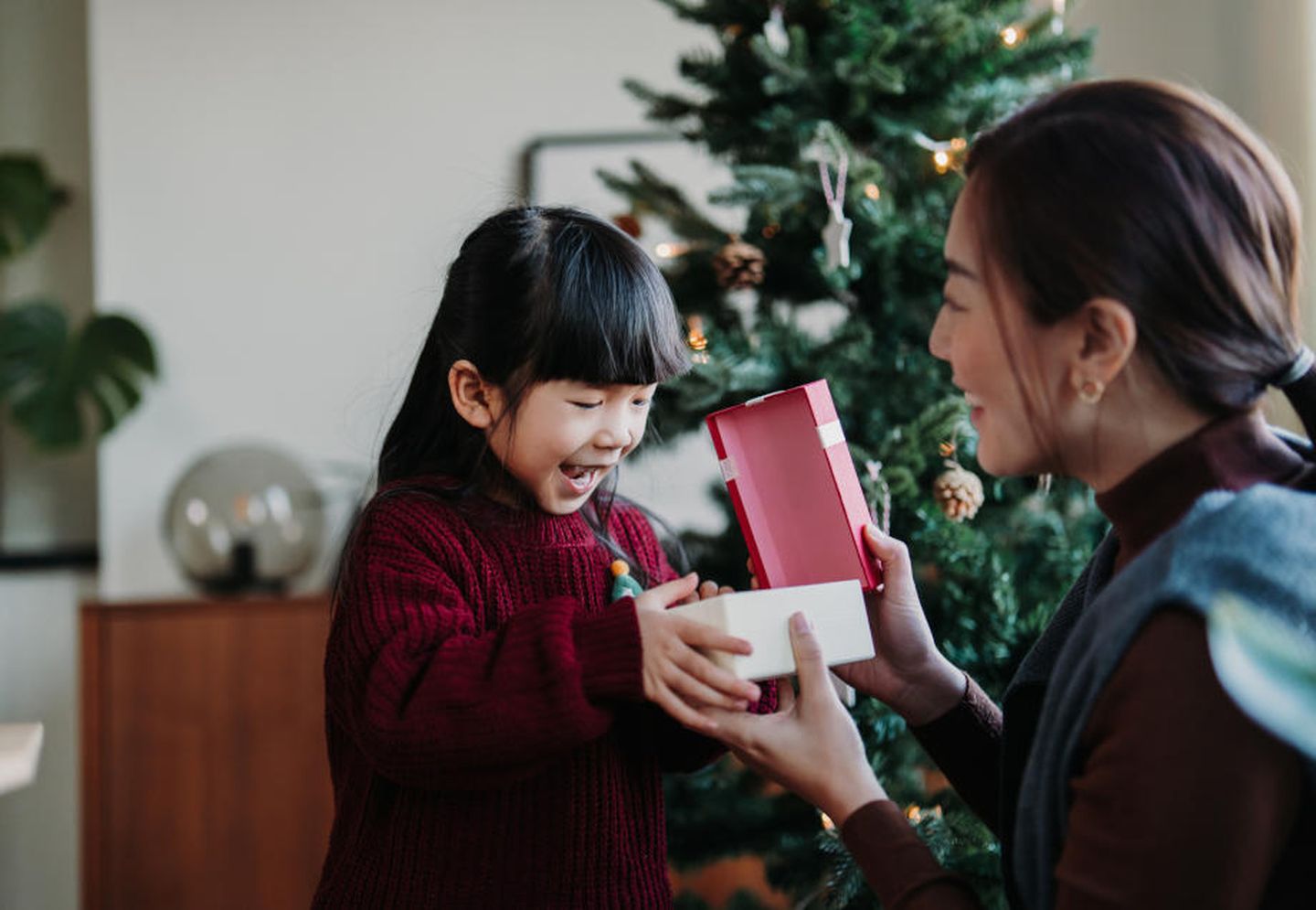 Mutter beschenkt Tochter vor einem Weihnachtsbaum. © d3sign / Moment via Getty Images