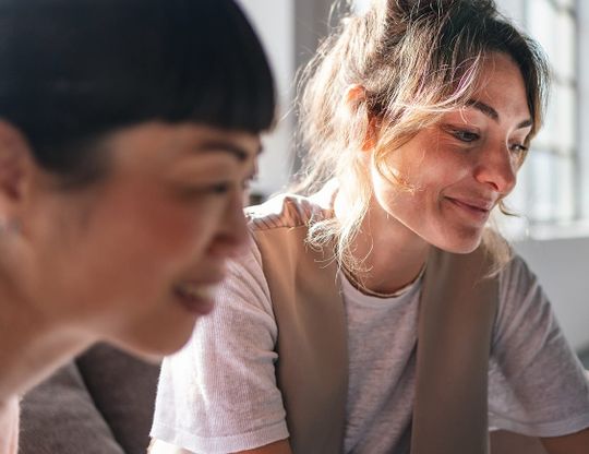 Two women in casual clothing share a warm moment in a sunlit interior one softly blurred in the foreground and one smiling in sharp focus behind
