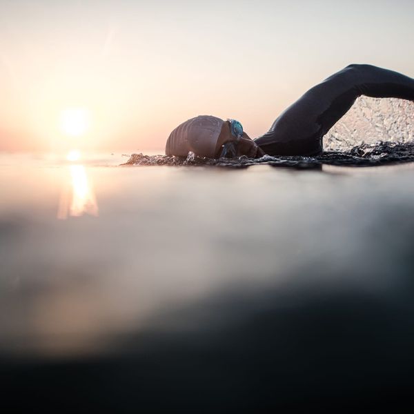 A swimmer in a wetsuit cap and goggles does front crawl in open water photographed at water level against a pastel sunrise sky