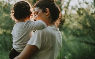 A person seen from behind carries a small child on their hip in a sunlit outdoor setting surrounded by green trees