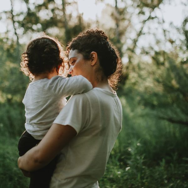 A person seen from behind carries a small child on their hip in a sunlit outdoor setting surrounded by green trees