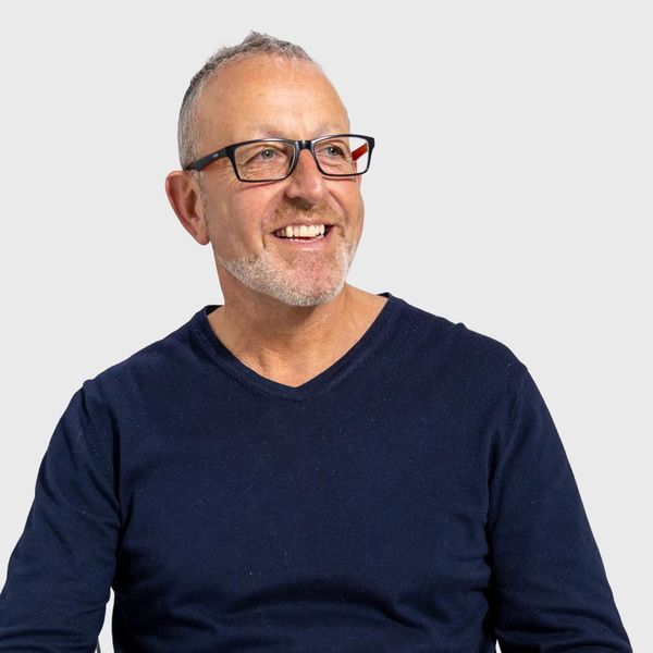 An older man with short grey hair and glasses wearing a navy v-neck jumper smiling broadly against a light grey background