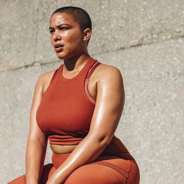 A woman with a shaved head wearing a rust-coloured ribbed sports top sits against a concrete wall skin glistening after exercise