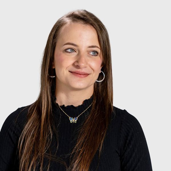 A young woman with long straight brown hair wearing a black ribbed top and a butterfly necklace smiling against a light grey background
