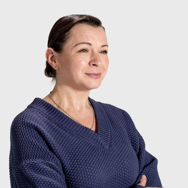 A woman with dark hair pulled back wearing a navy textured v-neck sweater with arms crossed looking slightly off-camera against a light grey background