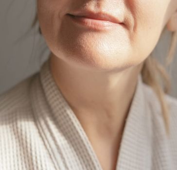 A close-up of the lower half of a woman's face chin and neck wearing a white textured robe with a gentle half-smile in soft natural light