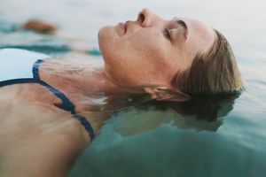 A woman in a blue swimsuit floats on her back in open water with eyes closed and a natural landscape visible in the soft background