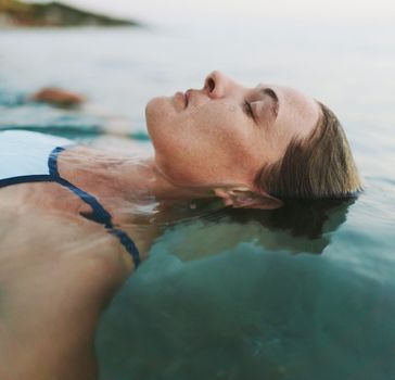A woman in a blue swimsuit floats on her back in open water with eyes closed and a natural landscape visible in the soft background
