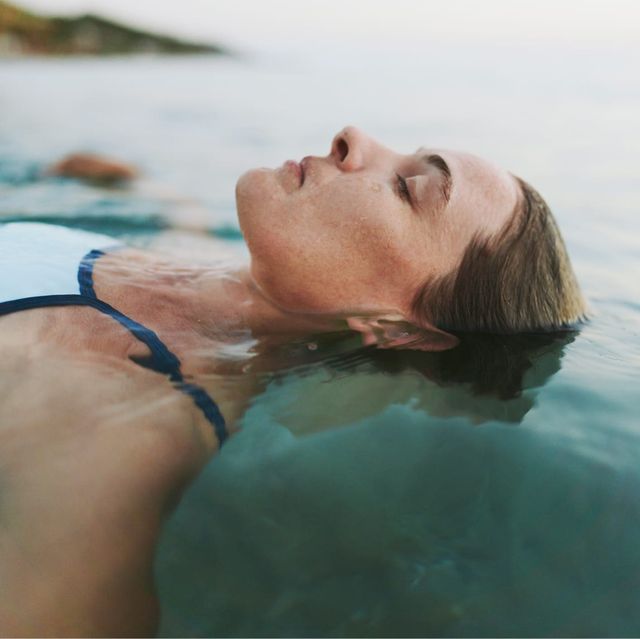 A woman in a blue swimsuit floats on her back in open water with eyes closed and a natural landscape visible in the soft background