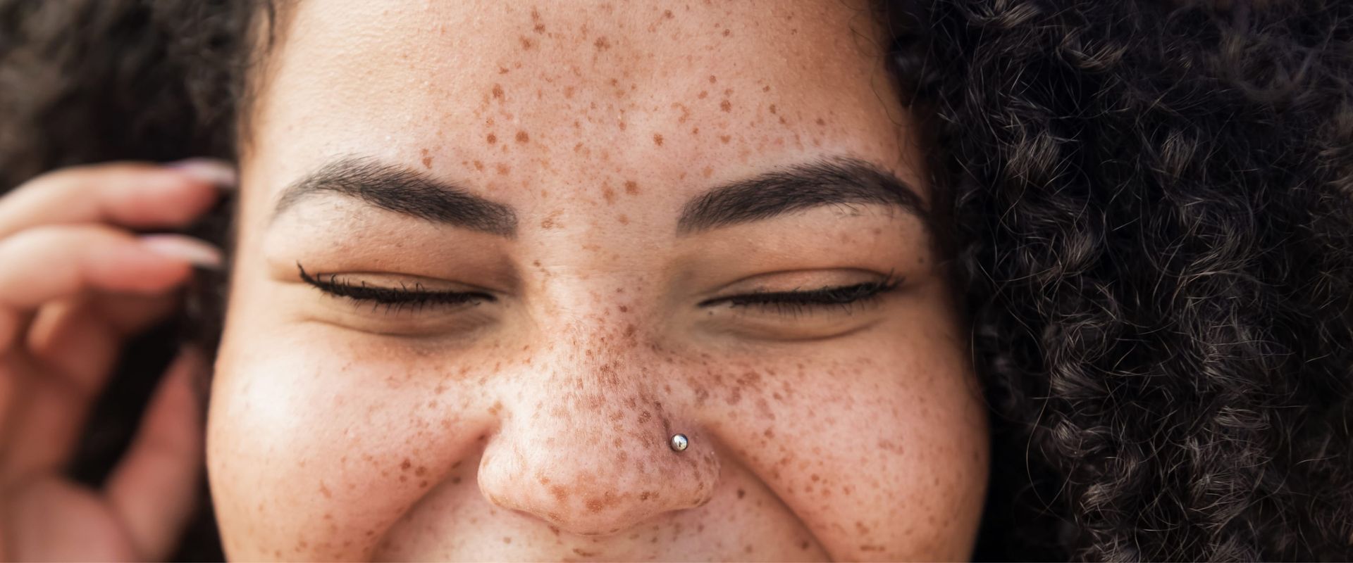 A close-up of a woman's face with freckles and curly dark hair with eyes closed in a smile and one hand raised near her temple