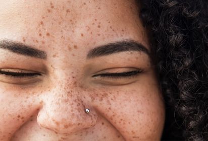 A close-up of a woman's face with freckles and curly dark hair with eyes closed in a smile and one hand raised near her temple