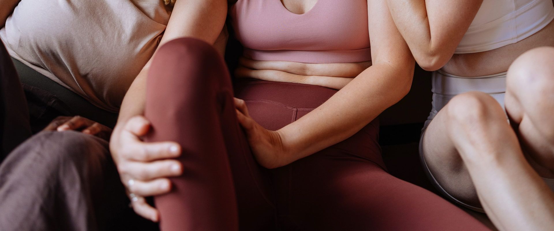 A woman in a pink sports bra and burgundy leggings sits surrounded and embraced by a group of friends in active wear