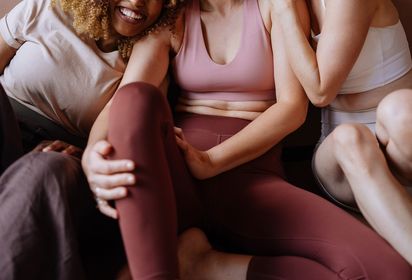 A woman in a pink sports bra and burgundy leggings sits surrounded and embraced by a group of friends in active wear
