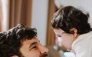A man with dark curly hair holds a young baby up to his face looking at the infant with a tender expression in a softly lit home setting