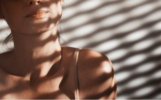 A close-up of a woman's neck collarbone and shoulder with a thin strap bathed in striped shadow patterns on a light background