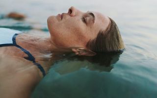 A woman in a blue swimsuit floats on her back in calm water with eyes closed and face in a relaxed expression