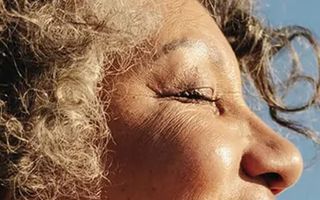 A close-up profile of an older woman's face with curly silver-grey hair eyes closed and face tilted upward toward a bright blue sky