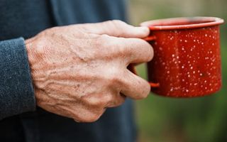 A close-up of an older person's weathered hands wrapped around a speckled red enamel mug outdoors