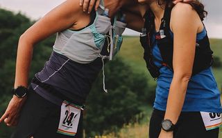 Two women wearing running vests and race bibs embrace on a trail course in an outdoor landscape sharing a moment of mutual support