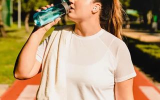 A woman in a white workout top drinks from a teal water bottle outdoors in bright sunlight with a towel draped over her shoulder