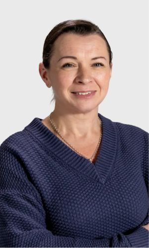 A woman with dark hair pulled back wearing a navy textured v-neck sweater arms partially crossed and smiling against a mint green background