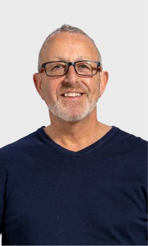 An older man with short grey hair and glasses wearing a navy v-neck top smiling warmly against a mint green background
