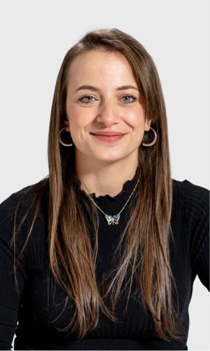 A young woman with long straight brown hair wearing a black ribbed top and a butterfly necklace smiling against a mint green background