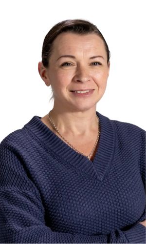 A woman with dark hair pulled back wearing a navy textured v-neck sweater arms partially crossed and smiling against a mint green background