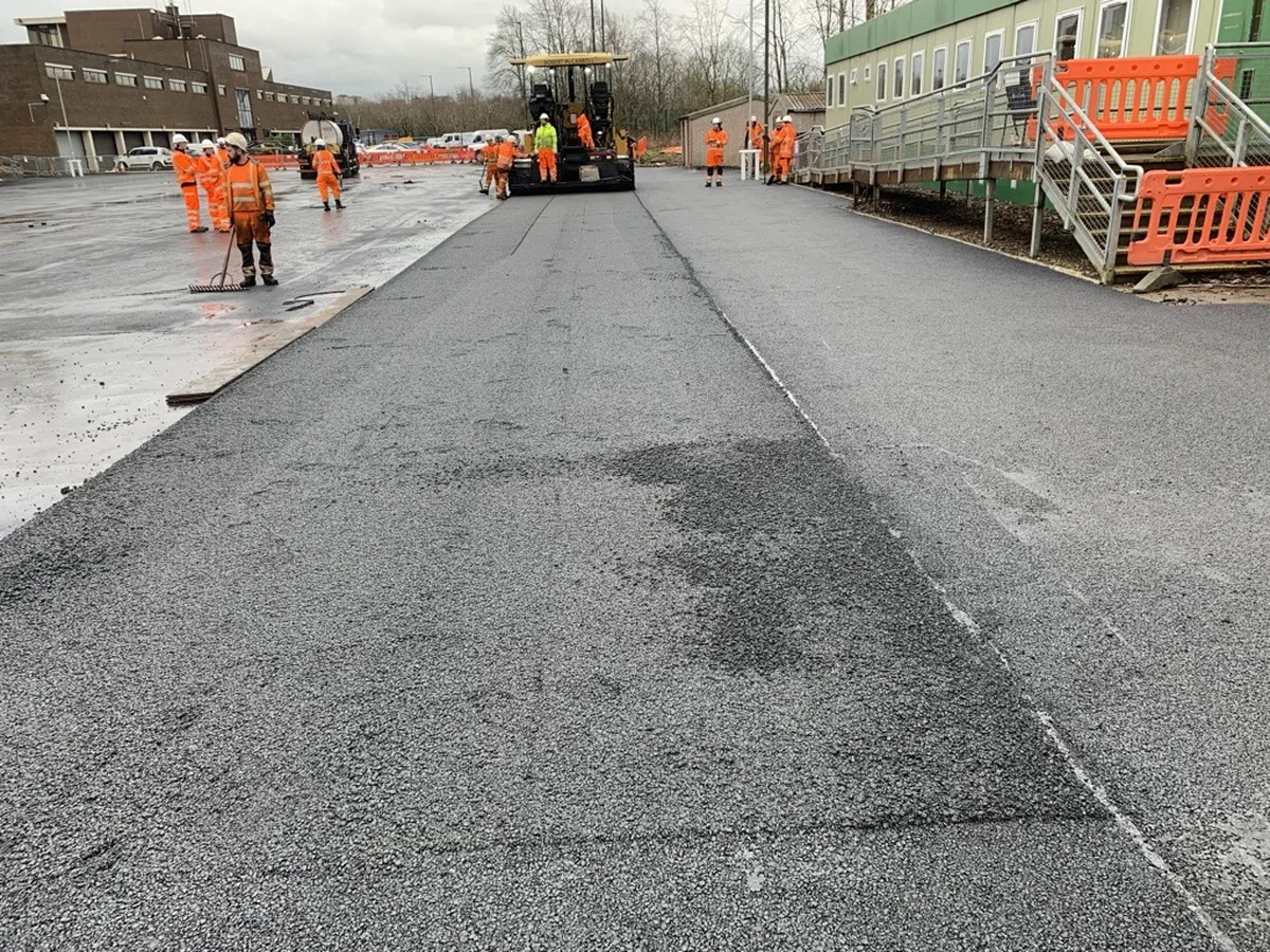 Workers in orange uniforms resurfacing a road with asphalt. Construction equipment and temporary buildings are visible nearby.