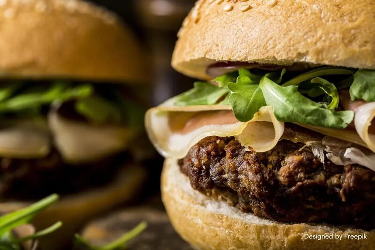 Close-up of a gourmet burger with a patty, arugula, and thinly sliced ham on a sesame bun. Another burger is blurred in the background.