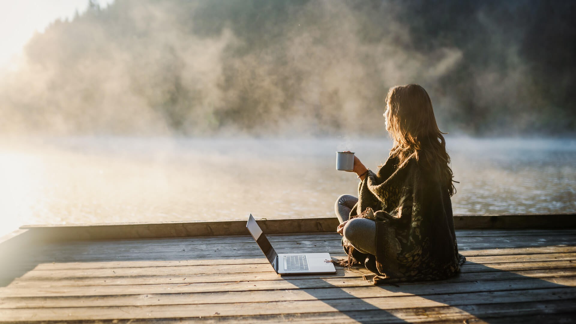 Eine Person sitzt auf einem hölzernen Steg mit einem Laptop und blickt auf einen See.