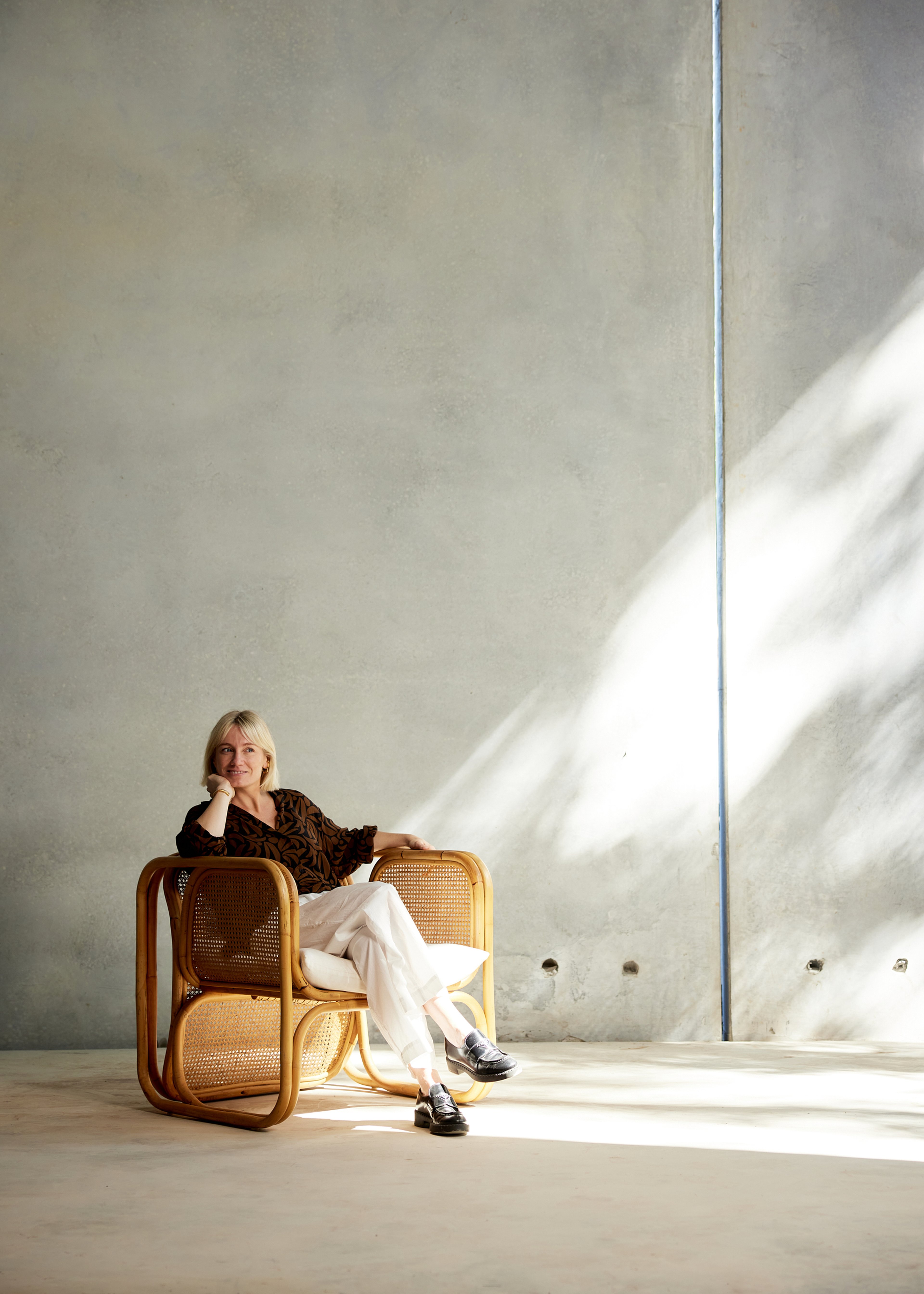 Susie owner of dayspa Comma sits on a woven chair in a sunlit room, wearing a patterned top and white pants, with a thoughtful expression.