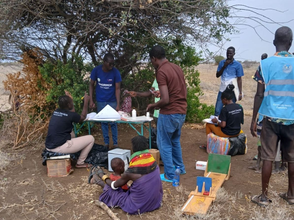 People gather around a makeshift outdoor station under a tree, attending to community activities and paperwork in a rural setting.