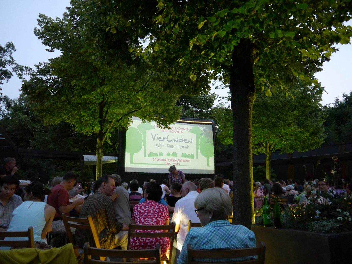 Publikum sitzt im Grünen vor einer Leinwand beim VierLinden Open-Air-Kino im Südpark Düsseldorf Metropol Düsseldorfer Filmkunstkinos GmbH