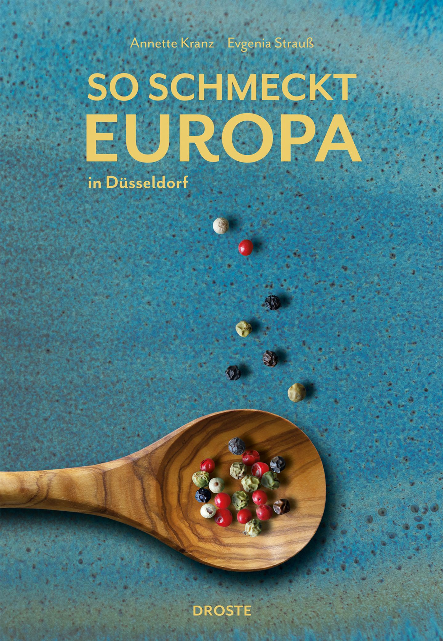 Das Cover eines Buches mit dem Titel "So Schmeckt Europa" zeigt einen Holzlöffel mit bunten Pfefferkörnern vor einem blauen Hintergrund.