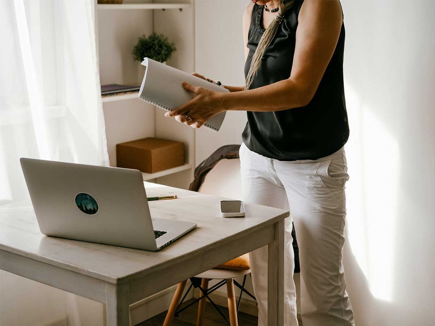 Eine Frau steht mit einem Block in der Hand vor einem Schreibtisch, auf dem sich ein Laptop und ein Handy befinden. © Fiordaliso / Moment via Getty Images