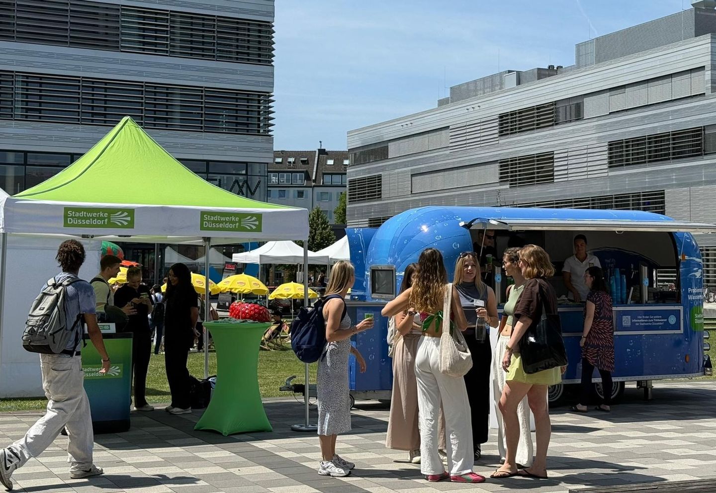 Outdoor-Veranstaltung mit Menschen in der Nähe eines Foodtrucks und grünen Zelten mit der Aufschrift „Stadtwerke Düsseldorf“, vor modernen Gebäuden.